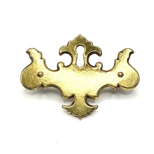 Könnte beinhalten: Ein goldfarbenes Metall-Schlüsselschild mit einer dekorativen, symmetrischen Form, geschwungenen Kanten und einer zentralen, vertikalen Schlüssellochöffnung. Das Stück verfügt über zwei kleine, kreisförmige Befestigungspunkte an den Seiten.