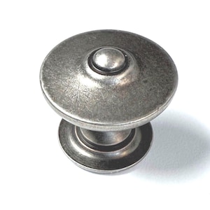 Puede incluir: Pomo de armario de estilo antiguo con diseño circular. El pomo presenta un botón central elevado y una base ancha y plana. El metal tiene un acabado gris desgastado, lo que sugiere un aspecto envejecido. Ideal para la restauración de muebles.