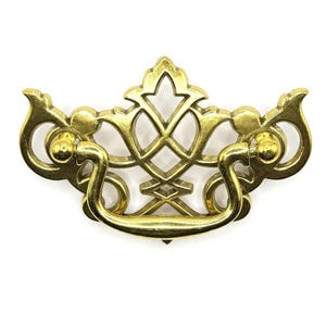 Puede incluir: Un tirador de cajón dorado con un diseño ornamentado. El tirador presenta un asa curva y elementos decorativos de filigrana. El herraje es de metal y está sobre un fondo blanco. El diseño general es simétrico y detallado.