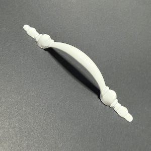 May include: A solid white cabinet handle featuring a smooth, arched center grip and decorative, rounded finials on both ends. The hardware has a glossy finish and a curved design, providing a functional grip for drawers or cabinet doors.