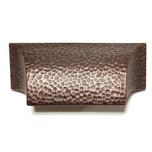 May include: A hammered copper cabinet pull with a rectangular shape and rounded edges. The pull has a textured surface and a warm, metallic finish.