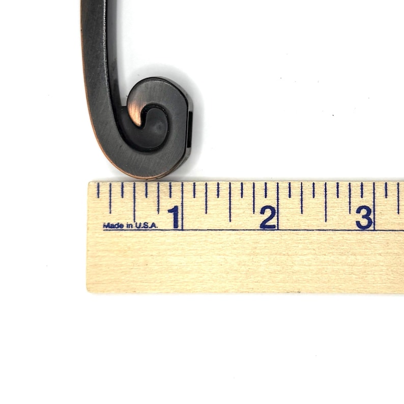 Pode incluir: Close-up de uma puxador de m&oacute;vel decorativo de metal com um design de rolagem. A puxador &eacute; marrom escuro com detalhes em cobre. Uma r&eacute;gua de madeira com marca&ccedil;&otilde;es em polegadas &eacute; colocada abaixo. A r&eacute;gua diz "Made in U.S.A."