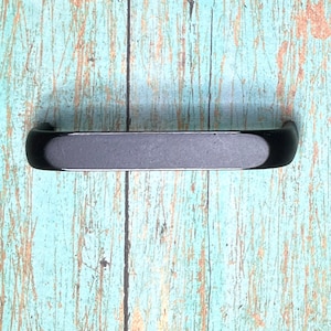 Puede incluir: Un tirador de cajón de metal negro mate con una forma rectangular alargada y extremos redondeados. El mango presenta un acabado oscuro, sólido y uniforme con un perfil curvo, diseñado para su fijación en muebles o cajones de armarios.