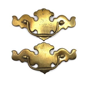 Op de afbeelding: Twee bijpassende messing ladehandgrepen met een decoratieve achterplaat met een geschulpte bovenrand en een gebogen, zwaaiende beugelgreep. Het metalen oppervlak vertoont een verweerde, verouderde patina met subtiele donkere vlekken.