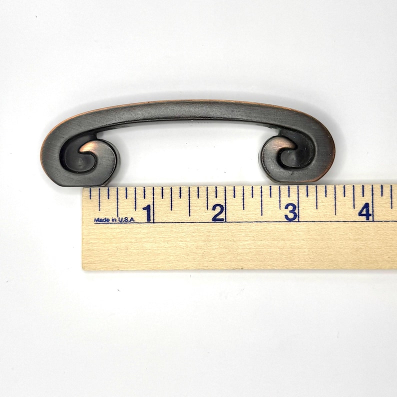 Pode incluir: Puxador de arm&aacute;rio cor de bronze com um design enrolado. O puxador tem aproximadamente 9 cm de comprimento, medido com uma r&eacute;gua de madeira. O puxador tem uma al&ccedil;a curva e extremidades decorativas enroladas. Feito nos EUA.
