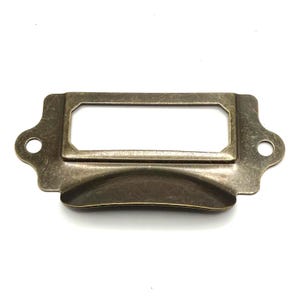 May include: A rectangular metal label holder with an antique bronze finish. The hardware features a central rectangular slot for a label, a curved handle grip along the bottom edge, and two circular mounting holes on the side extensions.
