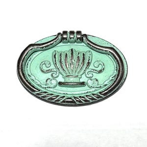 May include: Oval drawer pull in an antique style with a distressed finish. It has a light teal centre with an urn design and a dark silver frame. The design includes scrollwork and a leaf pattern. Suitable for furniture.