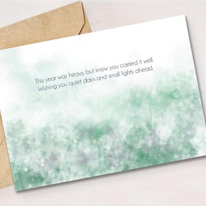 Puede incluir: Una tarjeta de felicitación con un diseño de acuarela en tonos verdes y blancos, acompañada de un sobre marrón. La tarjeta contiene el texto: "This year was heavy, but know you carried it well. Wishing you quiet days and small lights ahead."