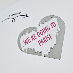 May include: A white greeting card with a heart-shaped cutout revealing the text "WE'RE GOING TO PARIS!" in pink. The card has a "scratch me!" prompt with an arrow. A pink envelope is visible in the background.