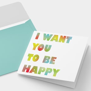 Könnte beinhalten: Eine weiße Grußkarte mit dem Text "I WANT YOU TO BE HAPPY" in Regenbogenfarben, neben einem offenen türkisfarbenen Umschlag. Die Karte ist gefaltet und leicht angewinkelt, wodurch die Botschaft deutlich wird.