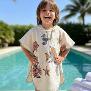 May include: A child wearing a cream-colored hooded beach towel with colorful sea creature designs. The towel features images of fish, seahorses, and starfish. The towel has green trim and is worn by a smiling child near a pool.