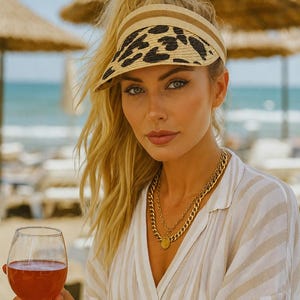 May include: A woman wearing a beige and black leopard print visor, gold necklaces, and a striped shirt, holding a glass of rosé wine. The scene is set on a sunny beach with straw umbrellas in the background.