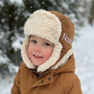 May include: A brown aviator hat with a cream-colored, fluffy lining and ear flaps. The hat has the name "Noah" embroidered in white on the side. The person in the image is wearing a brown jacket. The background is a snowy, outdoor scene.