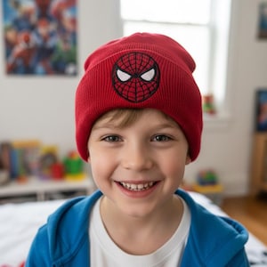 May include: A red knit beanie featuring a black and white embroidered Spider-Man emblem. The beanie is worn by a person wearing a blue hooded jacket and a white t-shirt. The background includes a poster and a window.