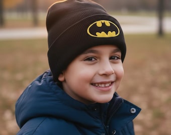 Könnte beinhalten: Eine schwarze Mütze mit einem gelben Batman-Logo. Die Mütze wird von einer Person getragen, die eine dunkelblaue Jacke trägt. Der Hintergrund ist verschwommen, mit Bäumen und Sonnenlicht.