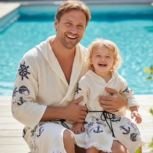 May include: Cream-colored hooded robes with navy blue nautical designs. The robes feature anchors, ship wheels, and other maritime symbols. The robes are made of a textured fabric and have a relaxed fit.
