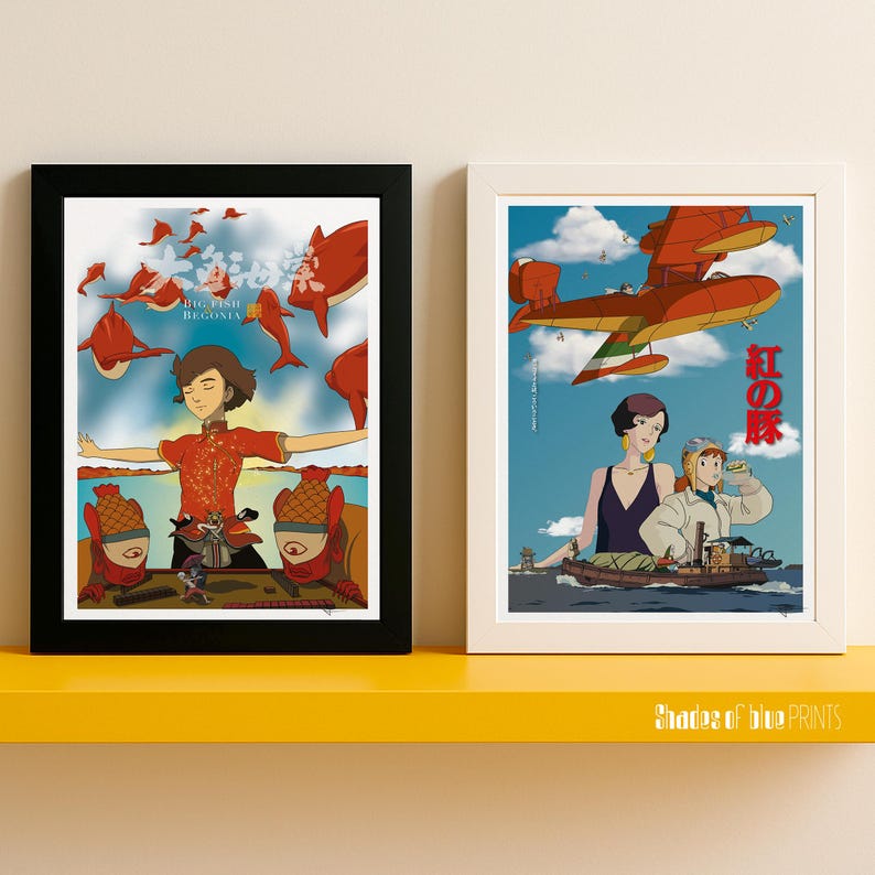 May include: Two framed art prints. The left print features a person in a red outfit with the text "Big Fish Begonia". The right print shows an orange seaplane and two women with Japanese text. Both prints are on a yellow shelf.