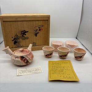 以下が含まれることがあります： 木箱、急須、湯呑み3客の日本の茶器セット。急須と湯呑みは、赤と黒のアクセントが付いた、テクスチャ加工されたオフホワイト仕上げです。木箱には日本語の文字が書かれています。