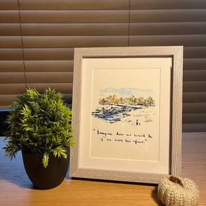 Könnte beinhalten: Ein gerahmtes Aquarellgemälde mit einem hellgrauen Rahmen, das eine Landschaft mit einem Fluss und Bäumen darstellt. Das Gemälde enthält das Zitat: „Stell dir vor, wie wir wären, wenn wir weniger Angst hätten.“ Eine kleine Topfpflanze und ein gestrickter Kürbis sind ebenfalls auf dem Tisch.