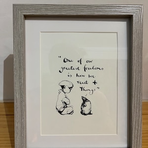 May include: A framed print with a light grey wooden frame. The artwork features a black and white illustration of two characters and handwritten text that reads, "One of our greatest freedoms is how we react to things."