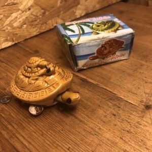 Puede incluir: Una figura de tortuga de cerámica en tonos beige y marrón, con una ranura para monedas. Junto a ella, una pequeña caja con una escena de playa tropical y la inscripción "Seashore Tortoise".