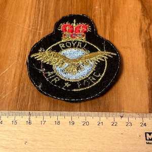 May include: A black patch with gold embroidery featuring a crown, eagle, and the words "ROYAL AIR FORCE". The patch has a blue and white circular design in the center. The patch is approximately 2 inches wide.
