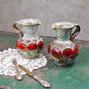 Può includere: Due brocche in ceramica con una smaltatura testurizzata verde chiaro maculata e una serie di medaglioni circolari rossi lucidi in rilievo attorno al corpo. Ogni brocca presenta un manico curvo e un beccuccio, accompagnate da una forchetta e un coltello.