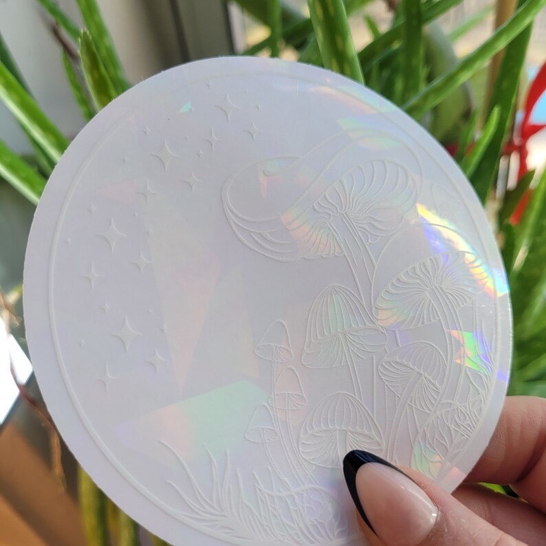 Puede incluir: Una pegatina blanca iridiscente con un dise&ntilde;o de setas y estrellas. La pegatina es redonda y tiene un acabado hologr&aacute;fico.