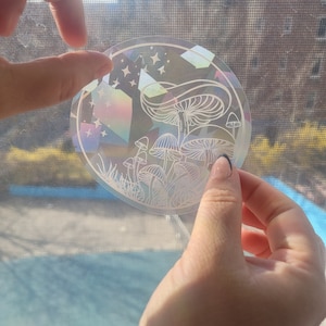 Puede incluir: Una pegatina hologr&aacute;fica con un contorno blanco de hongos y estrellas sobre un fondo iridiscente.
