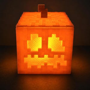 May include: A square, orange, pixelated jack-o'-lantern lamp with a blocky stem on top. The light emits a warm, glowing yellow hue through the cut-out eyes and jagged mouth, creating a distinct block-based aesthetic with visible 3D printed texture.