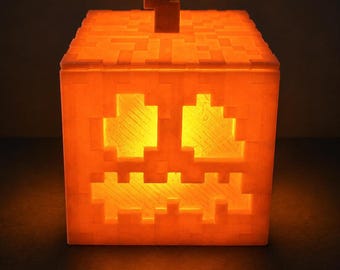 May include: A square, orange, pixelated jack-o'-lantern lamp with a blocky stem on top. The light emits a warm, glowing yellow hue through the cut-out eyes and jagged mouth, creating a distinct block-based aesthetic with visible 3D printed texture.