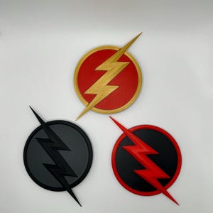 May include: Three Flash logo emblems. One is red with a gold lightning bolt, another is black with a black lightning bolt, and the third is black with a red lightning bolt. The emblems are circular.