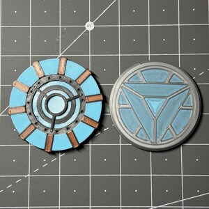 Puede incluir: Dos réplicas de reactor de arco de Iron Man impresas en 3D. Una es un reactor de arco de color azul y cobre con un diseño circular. La otra es un reactor de arco gris y azul con un diseño triangular.