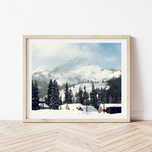 Könnte beinhalten: Ein gerahmtes Foto einer schneebedeckten Berglandschaft mit einer Gruppe von Nadelbäumen im Vordergrund. Der Berg ist schneebedeckt und der Himmel ist hellblau. Im Vordergrund befinden sich ein paar kleine Häuser.