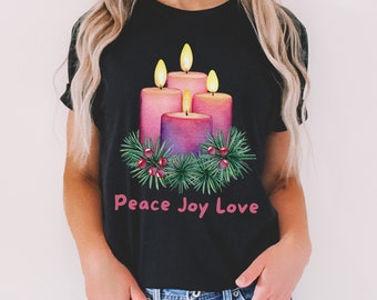 May include: Black t-shirt with a graphic of four purple and pink candles with flames, surrounded by a green wreath with red berries. The text "Peace Joy Love" is printed below the candles.
