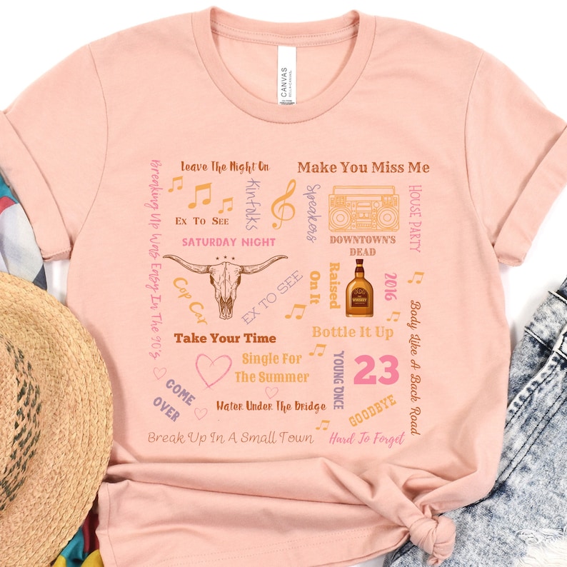 K&ouml;nnte beinhalten: Hellrosa T-Shirt mit einem Bullensch&auml;del-Motiv und Songtiteln. Das Design enth&auml;lt Noten und die Zahl "23". Das T-Shirt hat eine lockere Passform, ideal f&uuml;r den Alltag.