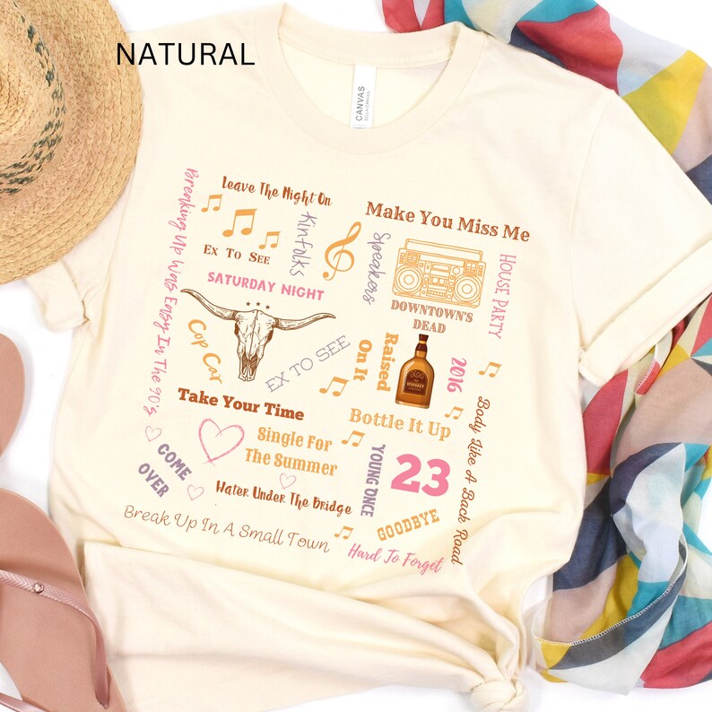 K&ouml;nnte beinhalten: Cremefarbenes T-Shirt mit verschiedenen Texten und Grafiken. Das Design umfasst einen Stiersch&auml;del, Noten, eine Boombox und S&auml;tze wie "Take Your Time" und "Make You Miss Me". Das Wort "NATURAL" ist oben aufgedruckt.