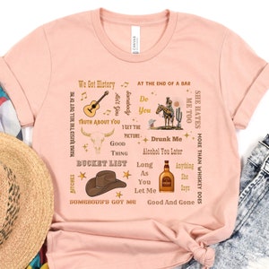 Puede incluir: Una camiseta rosa con una variedad de textos e imágenes impresos en ella. El texto incluye frases como "We Got History", "Truth About You", "Bucket List", "Somebody's Got Me", "Drunk Me", "Good And Gone", y "She Hates Me Too". Las imágenes incluyen una guitarra, una calavera, un sombrero de vaquero, una botella de whisky y un vaquero montando a caballo.