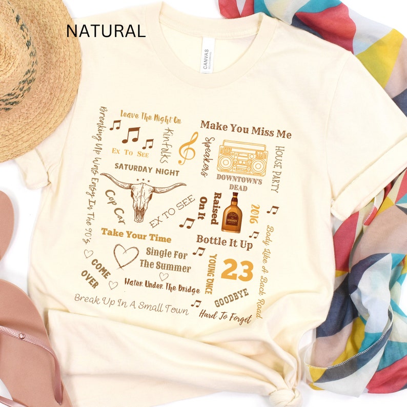 K&ouml;nnte beinhalten: Cremefarbenes T-Shirt mit einem grafischen Design mit Songtiteln, Noten und einem Longhorn-Sch&auml;del. Das Shirt enth&auml;lt den Text "23" und das Wort "NATURAL" oben.