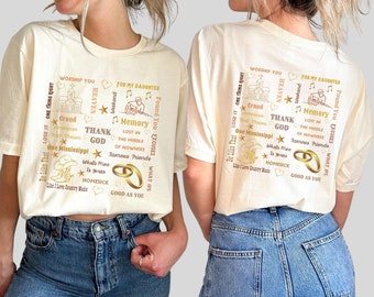 May include: Two white t-shirts with a gold design featuring words and phrases like "One Thing Right", "Worship You", "Grand Bad Dreams", "Thank God", "Lost in the Middle of Nowhere", "Famous Friends", "What's Mine is Yours", "Homesick", "Good as You", and "For My Daughter".