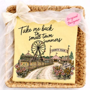 以下が含まれることがあります： 「Take me back to small town summers」の文字と、お祭りグラフィックが描かれた黄色のTシャツ。観覧車、お祭りの入り口、花などが描かれています。白いリボンが付いた、編みかごに入っています。