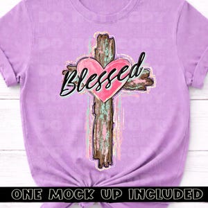 May include: A lilac-colored t-shirt featuring a graphic of a cross with a pink heart at the center. The word "Blessed" is written across the heart in black script. The cross has a rustic, wood-like appearance.