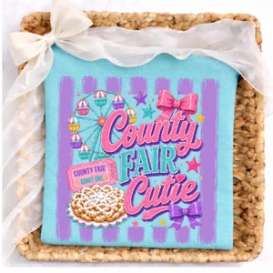 May include: A light blue t-shirt featuring a graphic print with purple vertical stripes, a Ferris wheel, a pink admission ticket labeled County Fair Admit One, a funnel cake, two pink bows, and the text County Fair Cutie in pink and blue.