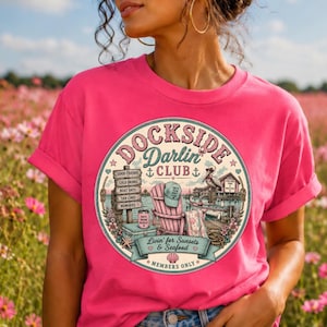 May include: A bright pink t-shirt featuring a circular graphic with the text DOCKSIDE DARLIN CLUB. The design includes a wooden sign, a beach chair with a cap, a pelican, a boat, and the text Livin for Sunsets & Seafood MEMBERS ONLY.