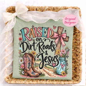 K&ouml;nnte beinhalten: Ein salbeigr&uuml;nes T-Shirt mit einem Grafikdruck und dem Text Raised on Dirt Roads & Jesus. Das Design zeigt ein Holzkreuz mit einer rosa Schleife, ein Paar braune Cowboystiefel mit Blumenmotiven, ein blaues Einmachglas mit Blumen und Herzen.