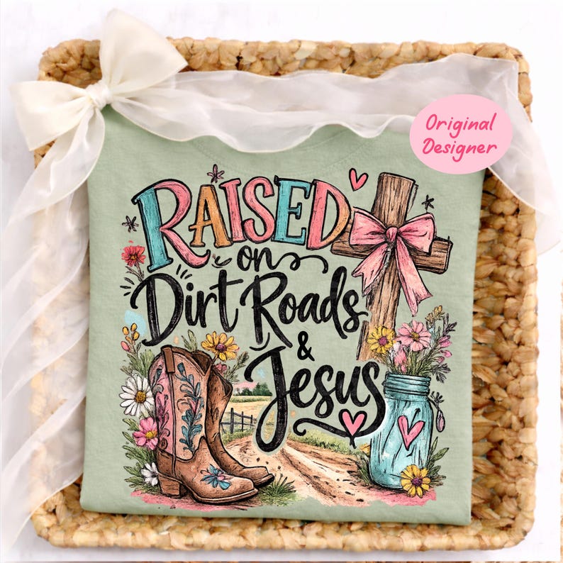 K&ouml;nnte beinhalten: Ein salbeigr&uuml;nes T-Shirt mit einem Grafikdruck und dem Text Raised on Dirt Roads & Jesus. Das Design zeigt ein Holzkreuz mit einer rosa Schleife, ein Paar braune Cowboystiefel mit Blumenmotiven, ein blaues Einmachglas mit Blumen und Herzen.