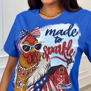 Op de afbeelding: Blauw T-shirt met een afbeelding van een haan met een zonnebril en een patriottische strik. De tekst "made to sparkle" staat boven de haan. Een rode schuur en een windmolen zijn ook afgebeeld.