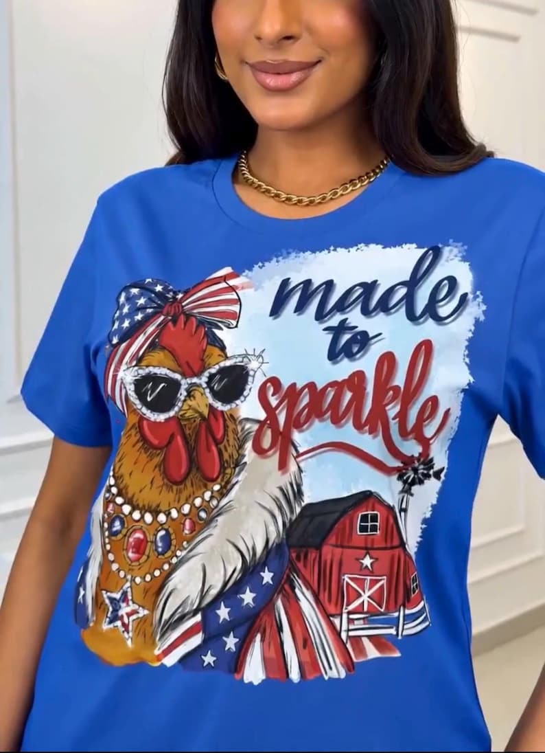 Op de afbeelding: Blauw T-shirt met een afbeelding van een haan met een zonnebril en een patriottische strik. De tekst "made to sparkle" staat boven de haan. Een rode schuur en een windmolen zijn ook afgebeeld.