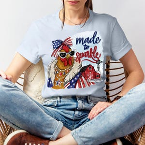 Op de afbeelding: Lichtblauw T-shirt met een cartoonkip met een zonnebril, een strik en een patriottische sjaal. De afbeelding bevat de tekst "made to sparkle" en een rode schuur met een windmolen. De kip draagt sieraden.