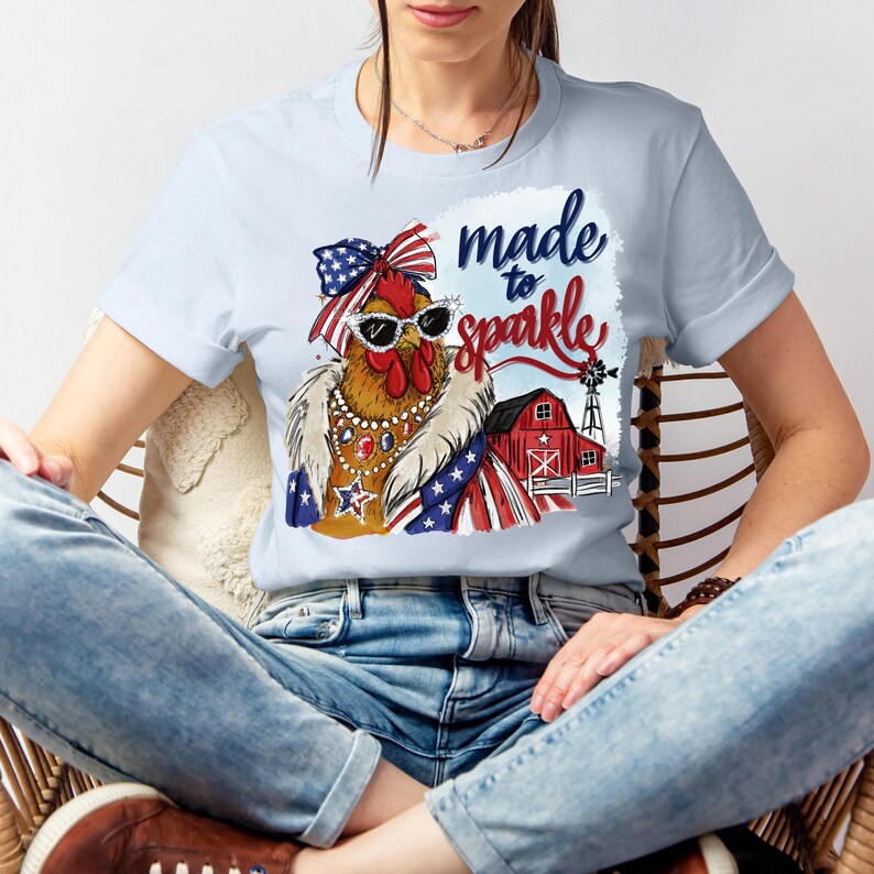Op de afbeelding: Lichtblauw T-shirt met een cartoonkip met een zonnebril, een strik en een patriottische sjaal. De afbeelding bevat de tekst "made to sparkle" en een rode schuur met een windmolen. De kip draagt sieraden.